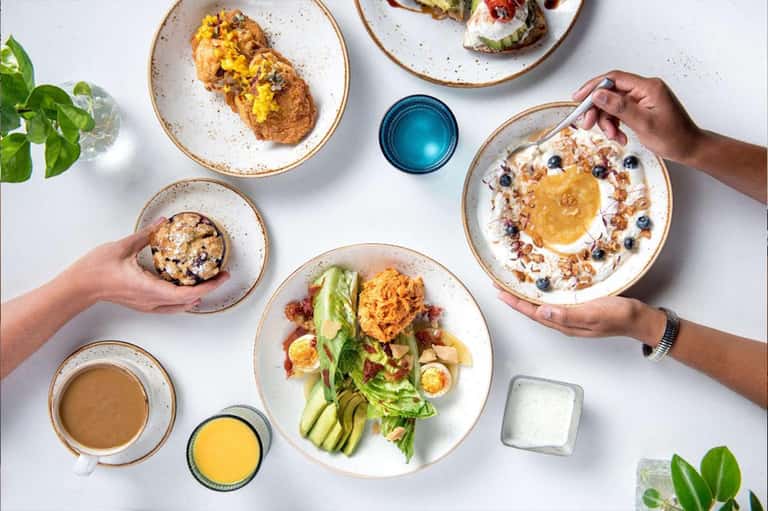 Overhead view of healthy breakfast dishes shared by multiple hands, including toast, yogurt, greens, and beverages on white plates