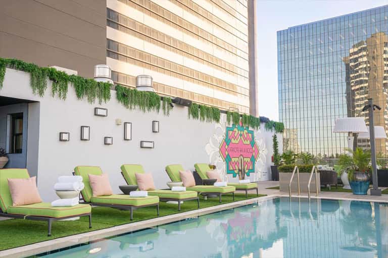 Resort pool with lime-green lounge chairs, pink cushions, decorative wall art, and modern hotel buildings