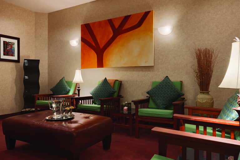 Lounge with tan textured wallpaper, red leather ottoman, green upholstered chairs, and abstract orange tree artwork