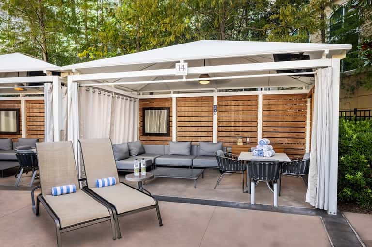 Outdoor poolside lounge area with white pergola, gray cushioned seating, wooden privacy fence, and striped chairs