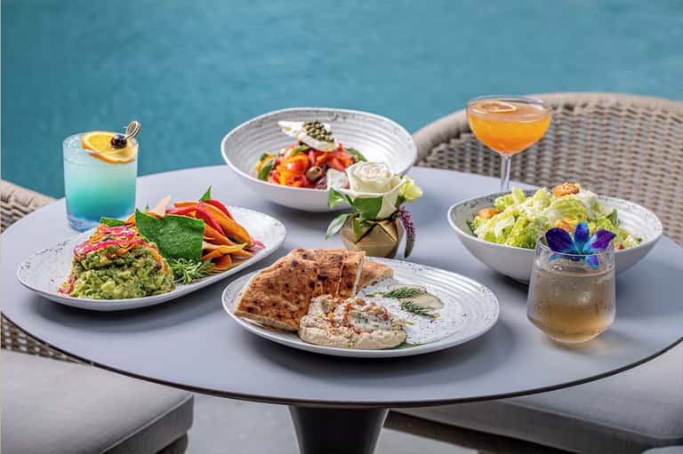 Poolside dining table with gourmet plated dishes, cocktails, and colorful edible flowers on white dinnerware