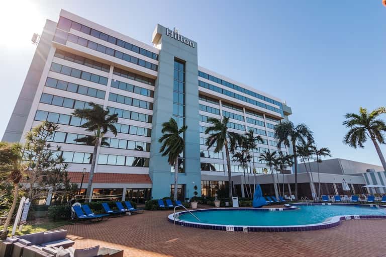 Modern Hilton hotel exterior with pool, blue loungers, palm trees, and multi-story building facade