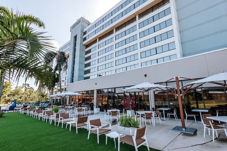 Modern hotel exterior with palm trees and ground-level outdoor patio dining area with umbrellas