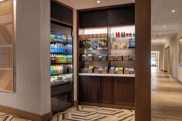 Modern hotel snack bar with refrigerated beverage display and woven basket storage shelving