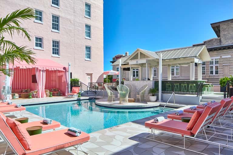 Resort pool area with pink lounge chairs, white pergola, palm trees, and multi-story hotel building