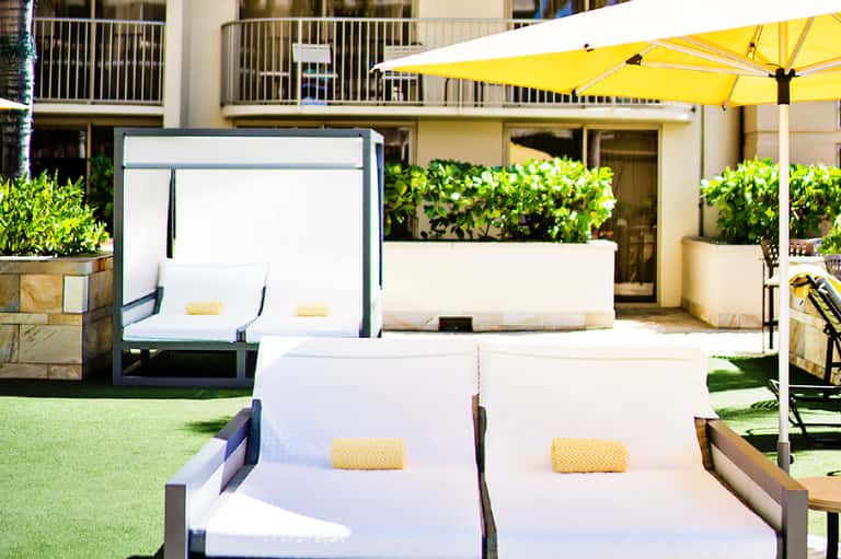 Modern poolside lounge with white sunbeds, yellow umbrella, manicured lawn, and building in background