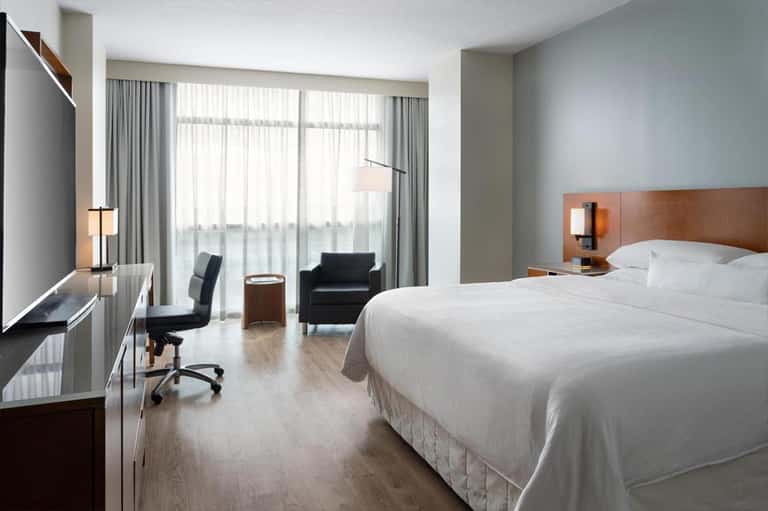 Modern hotel bedroom with king bed, work desk, armchair, and floor-to-ceiling windows