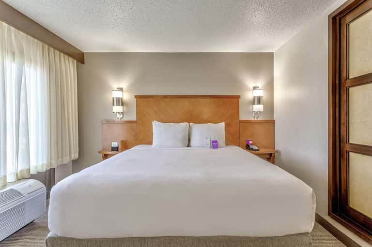 Modern hotel bedroom with wood headboard, white bedding, elegant wall sconces, and bright window light