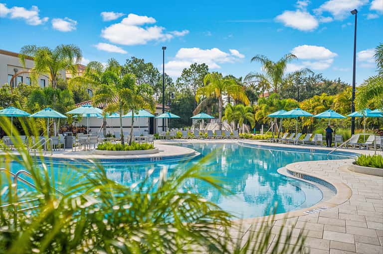 Resort pool area with blue umbrellas, lounge chairs, palm trees, and clear sunny sky