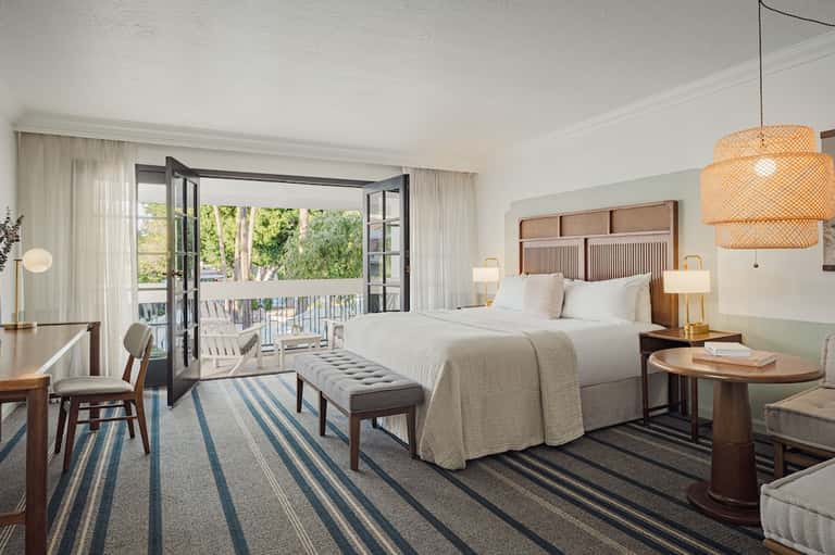 Bright hotel bedroom with king bed, balcony doors, striped rug, and views of nature