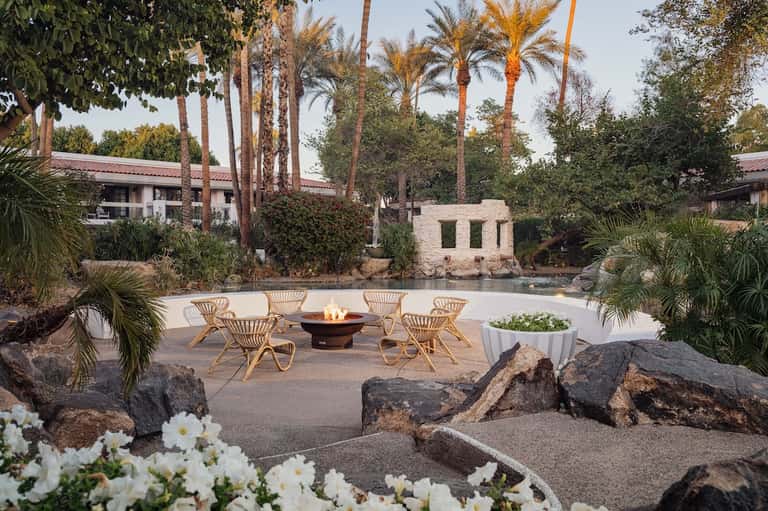 Outdoor fire pit lounge with woven chairs, palm trees, water feature, and tropical landscaping