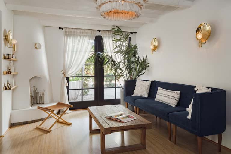 Bright living room with white walls, navy sofa, wooden coffee table, hanging light fixture, potted plant, and glass doors with curtains