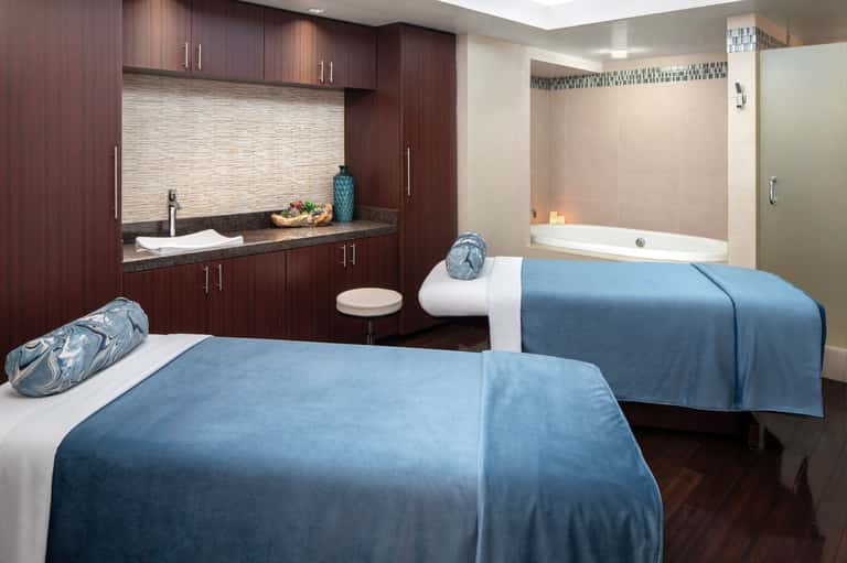 Spa treatment room with two massage beds, dark wood cabinetry, and soaking tub