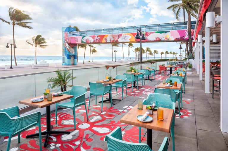 Beachfront dining patio with turquoise chairs, colorful floral canopy, and ocean views