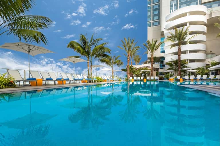 Resort pool with crystal blue water, palm trees, white umbrellas, and modern curved architecture