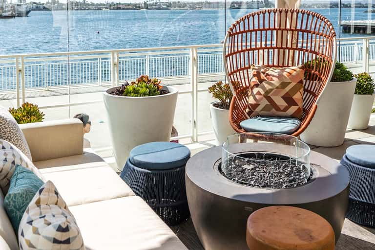 Waterfront balcony lounge with woven chair, fire pit, potted plants, and bay views