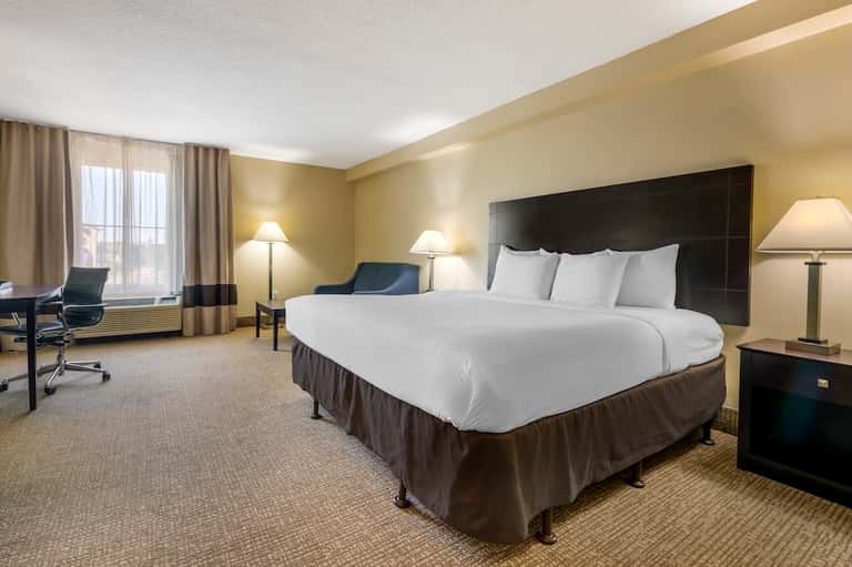 Spacious hotel bedroom with queen bed, black headboard, beige walls, work desk, and window with city views.