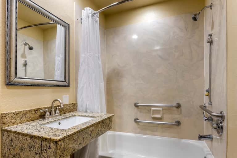 Hotel bathroom with granite countertop sink, mirror, white shower curtain, and tub with grab bars