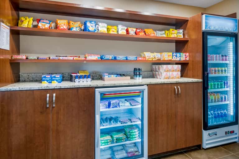 Hotel snack bar with wooden cabinetry, open shelving displaying candies and snacks, and beverage cooler.