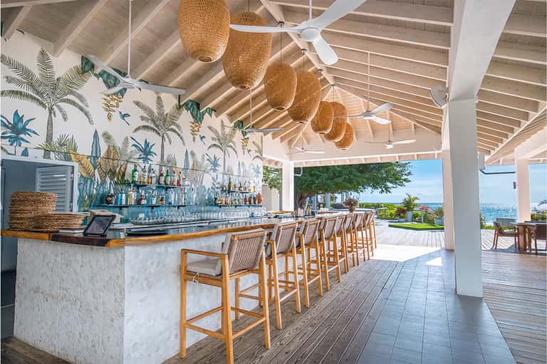 Tropical beach bar with white columns, woven pendant lights, wooden stools, and ocean views