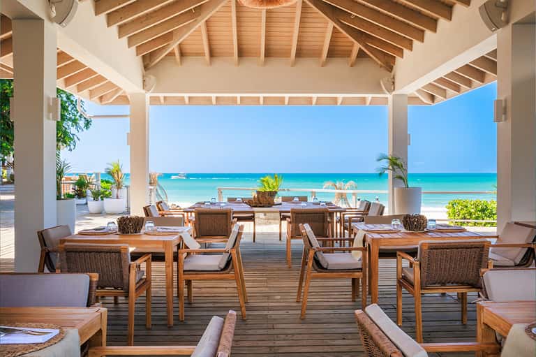 Beachfront dining pavilion with wooden tables, rattan chairs, and clear ocean view under white roof