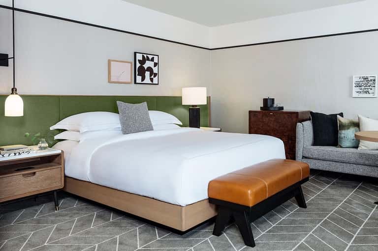 Contemporary hotel bedroom with green headboard, white bedding, wood furniture, and modern artwork.