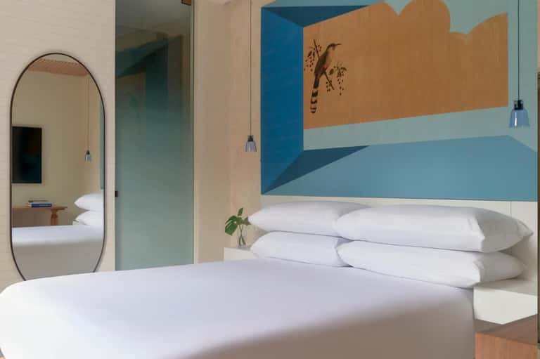 Modern hotel bedroom with white bedding, arched mirror, blue and tan wall mural, and pendant lighting