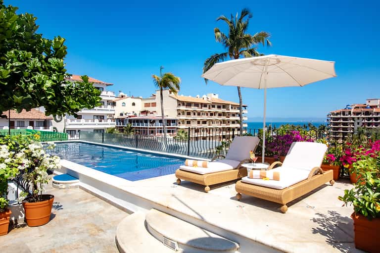 Mediterranean resort pool with white umbrella, lounge chairs, sea view, and colorful blooming flowers