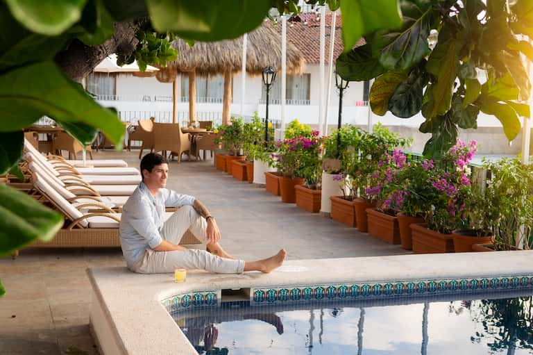 Man in white linen sitting by tropical pool with palm trees, thatched-roof structures, and white colonial-style building