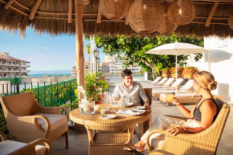 Couple dining at a round table on a rooftop terrace with ocean views and lounge chairs in background