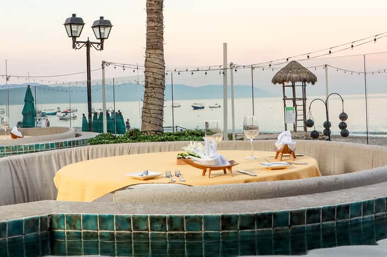 Oceanfront dining setup with yellow tablecloth, palm tree, and sea view beyond glass railing