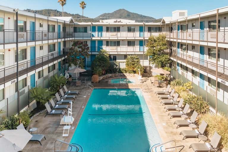 Aerial view of hotel courtyard with two pools, palm trees, and surrounding multi-story blue and white buildings