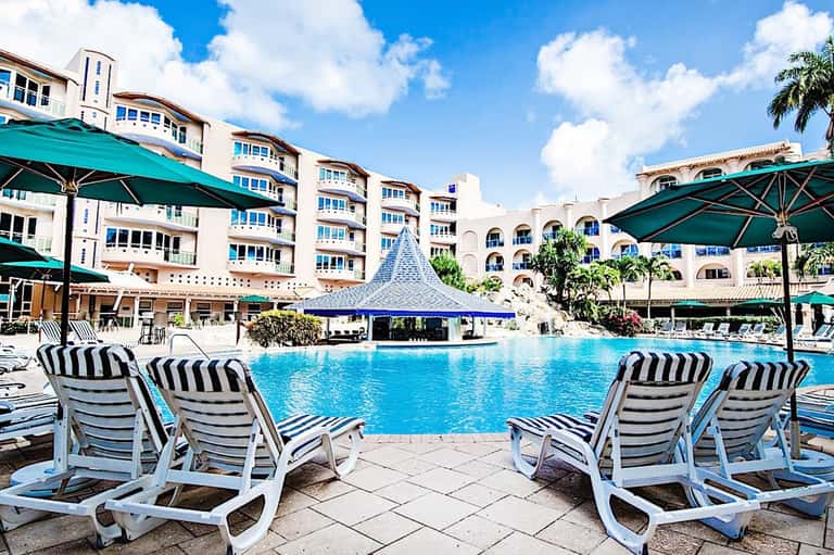 Resort pool with striped lounge chairs, green umbrellas, and multi-story hotel buildings