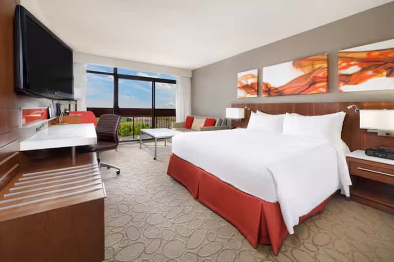 Hotel guest room with queen bed, flat-screen TV, desk, and balcony overlooking landscape