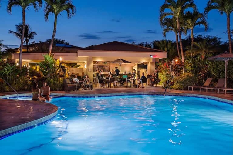 Resort pool at night with illuminated pavilion, palm trees, and guests socializing