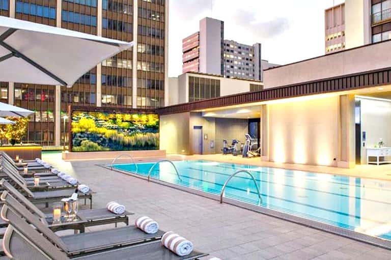 Modern indoor swimming pool with loungers, green living wall, and city skyscrapers visible through floor-to-ceiling windows