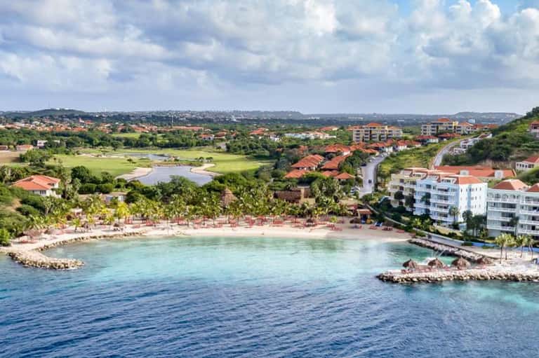 Coastal beachfront resort with pink sand beach, turquoise water, residential buildings, and lush green hills