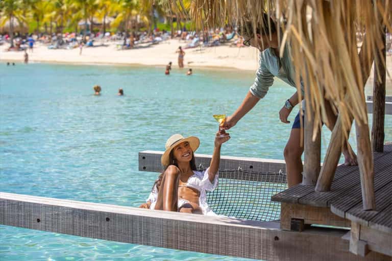 Woman in white sun hat enjoying drink at beachfront cabana with ocean and palm trees in background