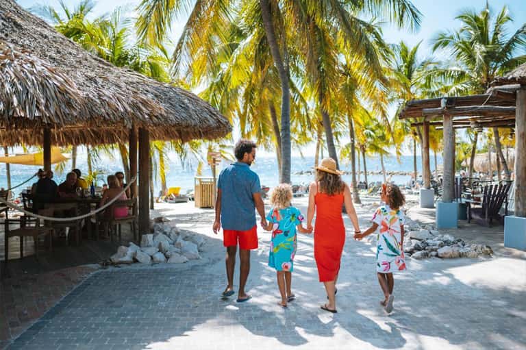 Family walking on sandy beach with thatched-roof cabanas, palm trees, and beachfront resort