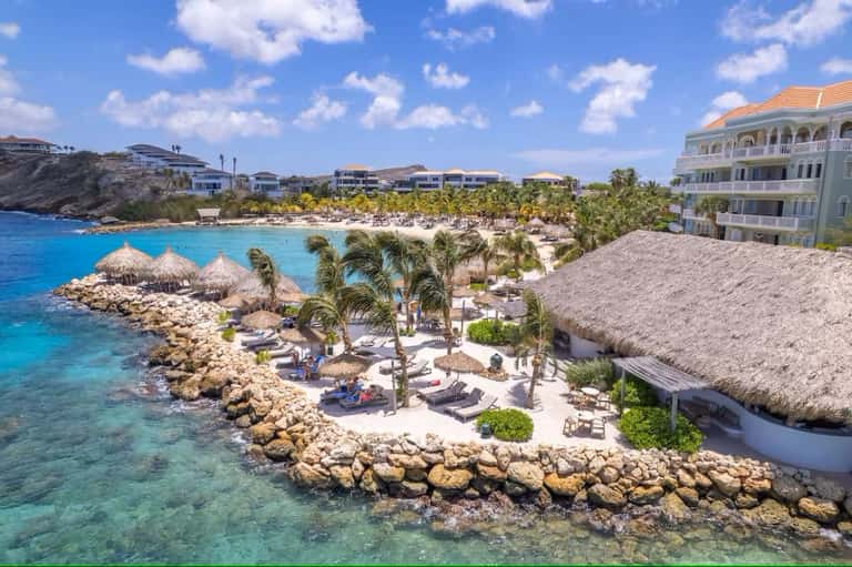 Beachfront resort with blue ocean, thatched cabanas, palm trees, and multi-story building