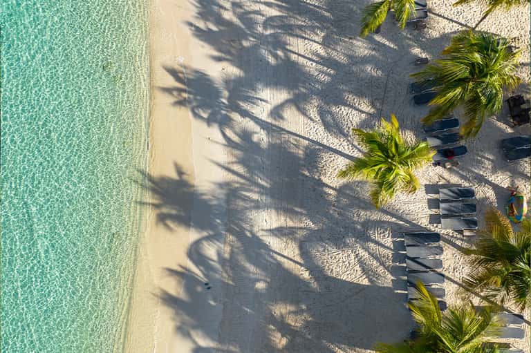 Aerial view of pristine turquoise ocean waters and sandy beach with palm tree shadows and lounge chairs