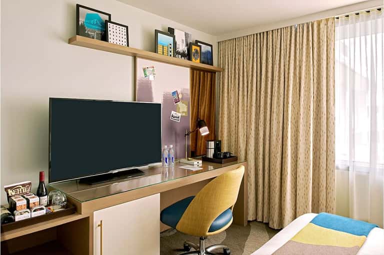 Hotel room workspace with flat-screen TV, wooden desk, yellow chair, and large window with natural light