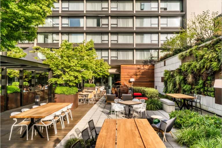 Outdoor courtyard with wooden dining tables, lush greenery, and covered lounge area