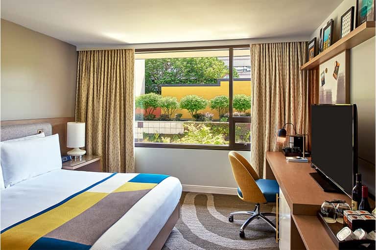 Contemporary hotel bedroom with colorful bedding, work desk, window overlooking garden with yellow accent wall