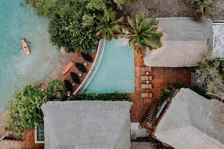 Aerial view of resort pool, wooden deck, palm trees, and turquoise water surrounded by buildings