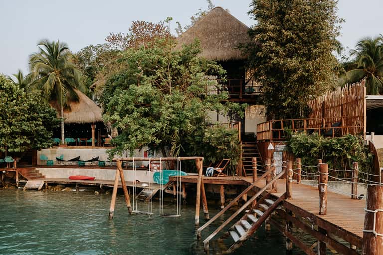 Tropical waterfront resort with thatched-roof buildings, wooden docks, and lush vegetation