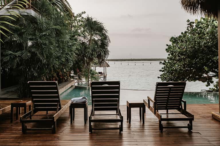 Waterfront lounge chairs on wooden deck overlooking bay with palm vegetation