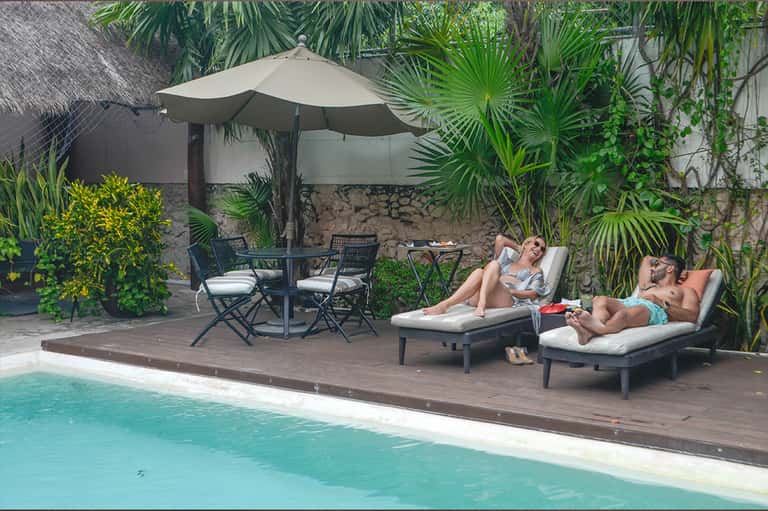 Tropical poolside lounge with umbrellas, wooden deck, palm plants, and guests relaxing on loungers by pool