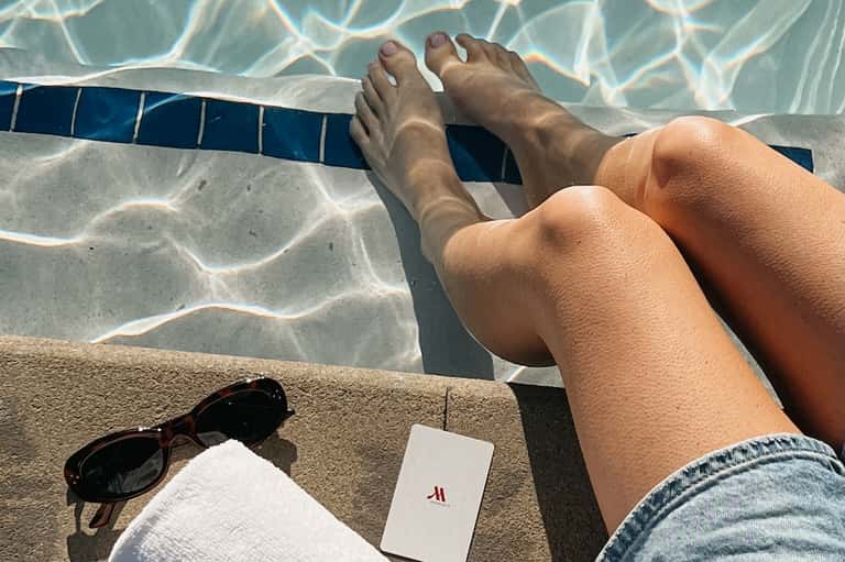 First-person perspective of feet dangling over pool edge with crystal-clear water and blue tile visible