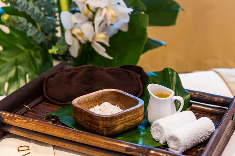 Spa amenities on wooden tray with white towels, wooden bowl with salt, oil pitcher, and white flowers